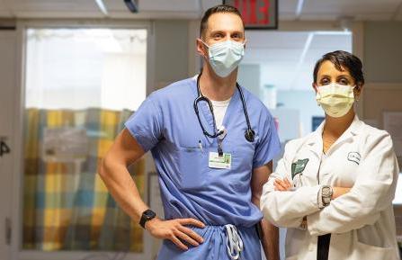 年轻的印度女子穿着白色实验室大褂，戴着外科口罩，双臂交叉，站在一个高个子白人男子旁边，他穿着手术服，手放在臀部，戴着听诊器, 在重症监护室前的走廊里